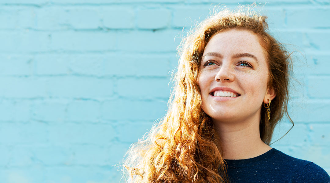 A girl with long curly hair against a blue brick background looking up and smiling